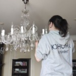 Співробітниця у фірмовому одязі компанії Юнона акуратно очищує кришталеву люстру під час генерального прибирання.