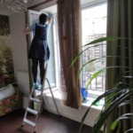 Співробітниця клінінгової компанії Юнона на стрем'янці ретельно миє високе вікно та пластикову раму в квартирі.